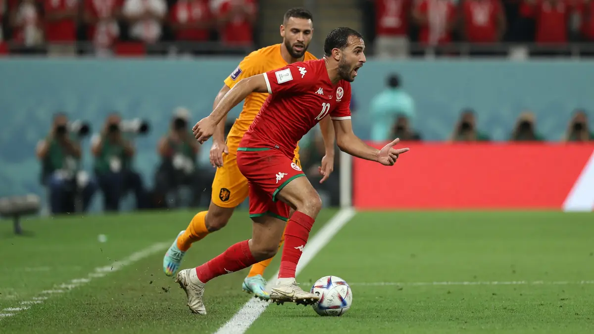 Tight sideline action frame of a left-sided attacker driving with the ball under stadium lights, captured with shallow depth of field and the crowd blurred behind the touchline